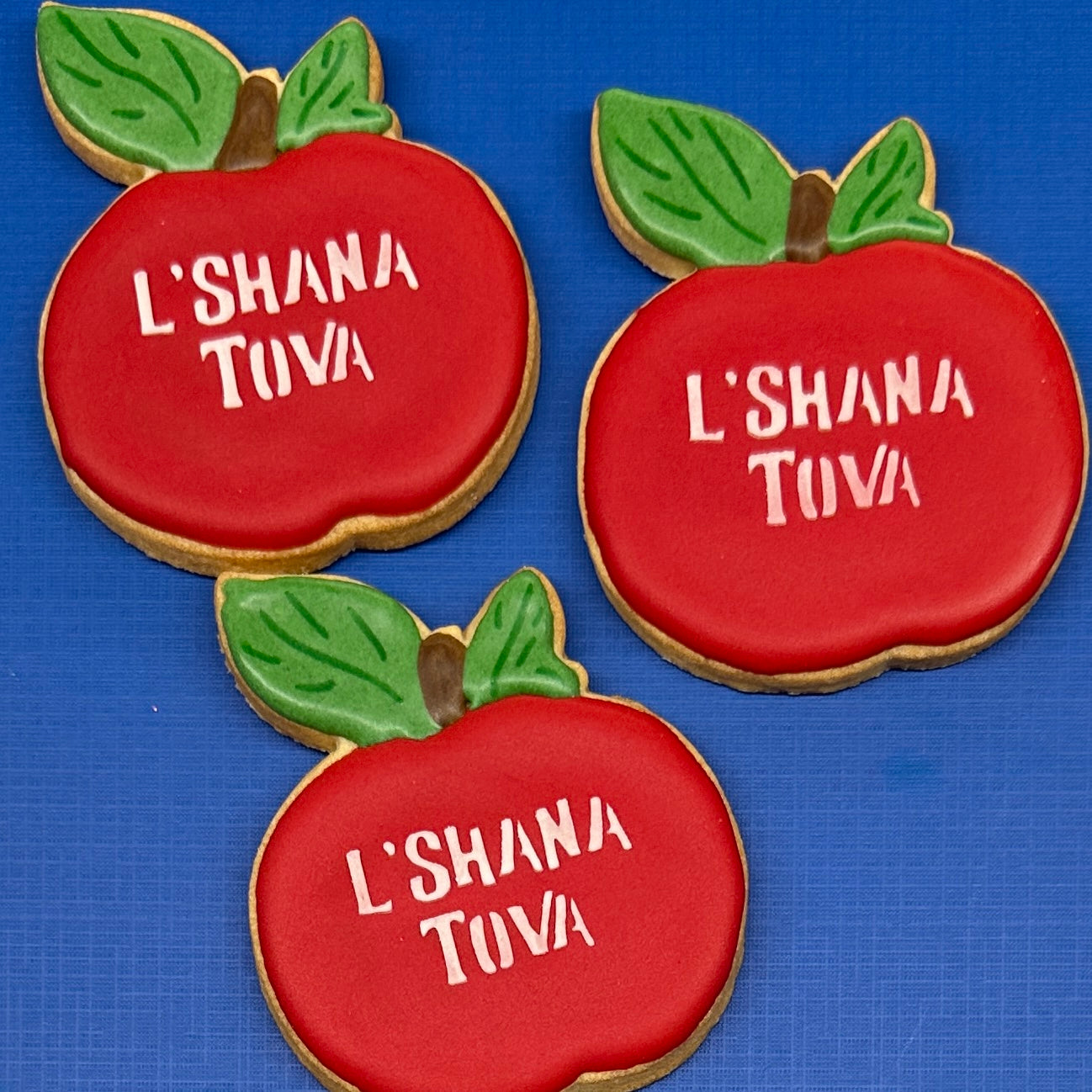 Apple-shaped sugar cookies decorated with red icing, green leaves, and white icing text reading 'L'SHANA TOVA'.