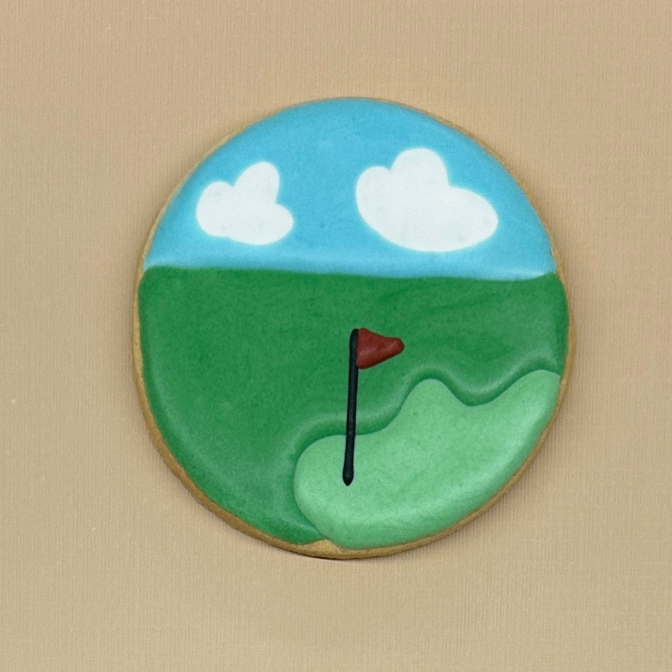 A round sugar cookie decorated with golf course scenery, featuring a green fairway, a blue sky with white clouds, and a red flag, set against a neutral beige background.