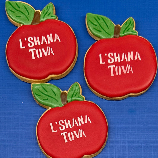 Apple-shaped sugar cookies decorated with red icing, green leaves, and white icing text reading 'L'SHANA TOVA'.