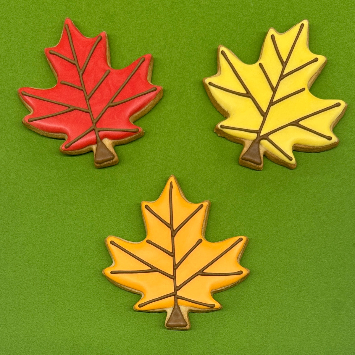 Three maple leaf-shaped cookies decorated with autumn colors, arranged on a solid green background. One cookie is red, and two are yellow with brown stems and vein details.