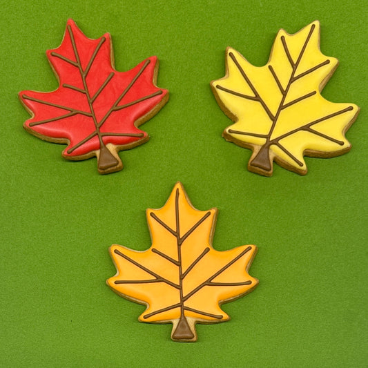 Three maple leaf-shaped cookies decorated with autumn colors, arranged on a solid green background. One cookie is red, and two are yellow with brown stems and vein details.