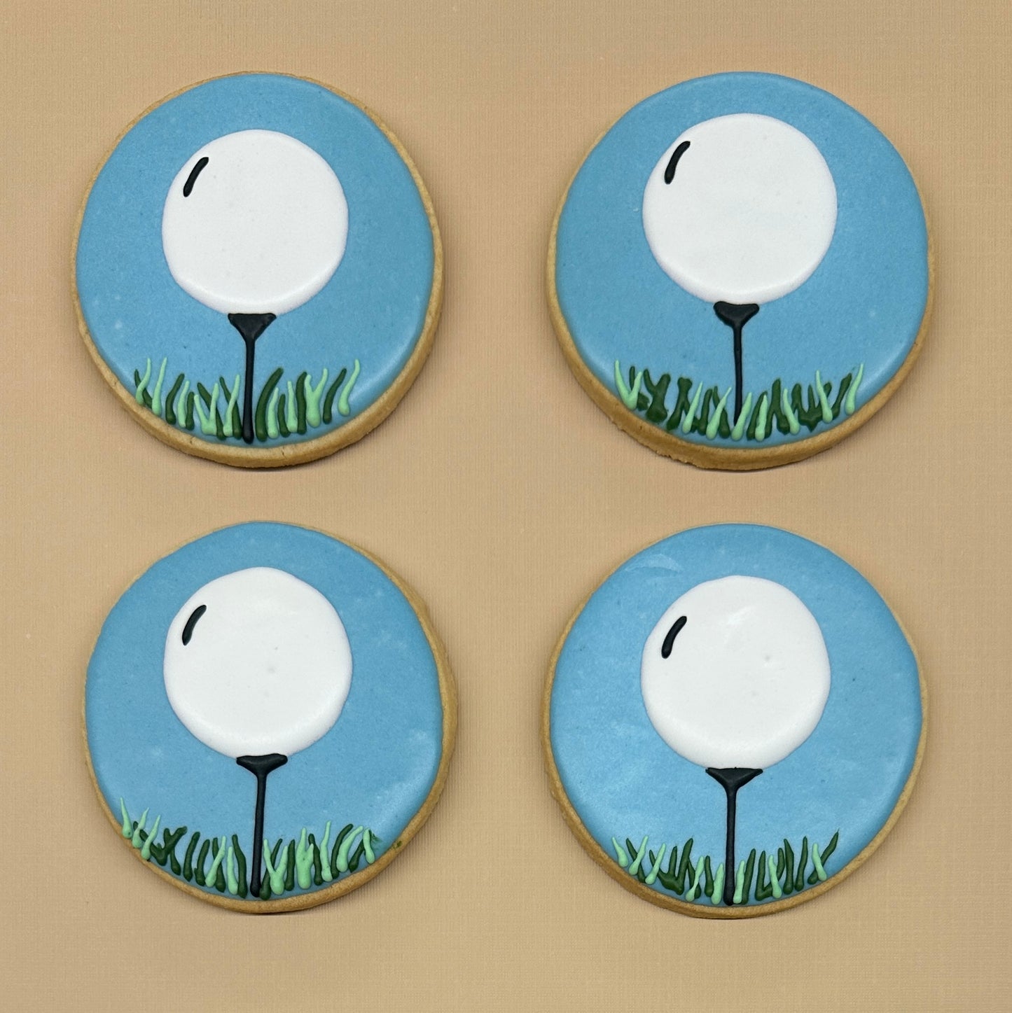 Four round sugar cookies decorated with golf-themed icing, featuring a white golf ball on a black stand with green grass against a light blue background.