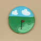 A round sugar cookie decorated with golf course scenery, featuring a green fairway, a blue sky with white clouds, and a red flag, set against a neutral beige background.